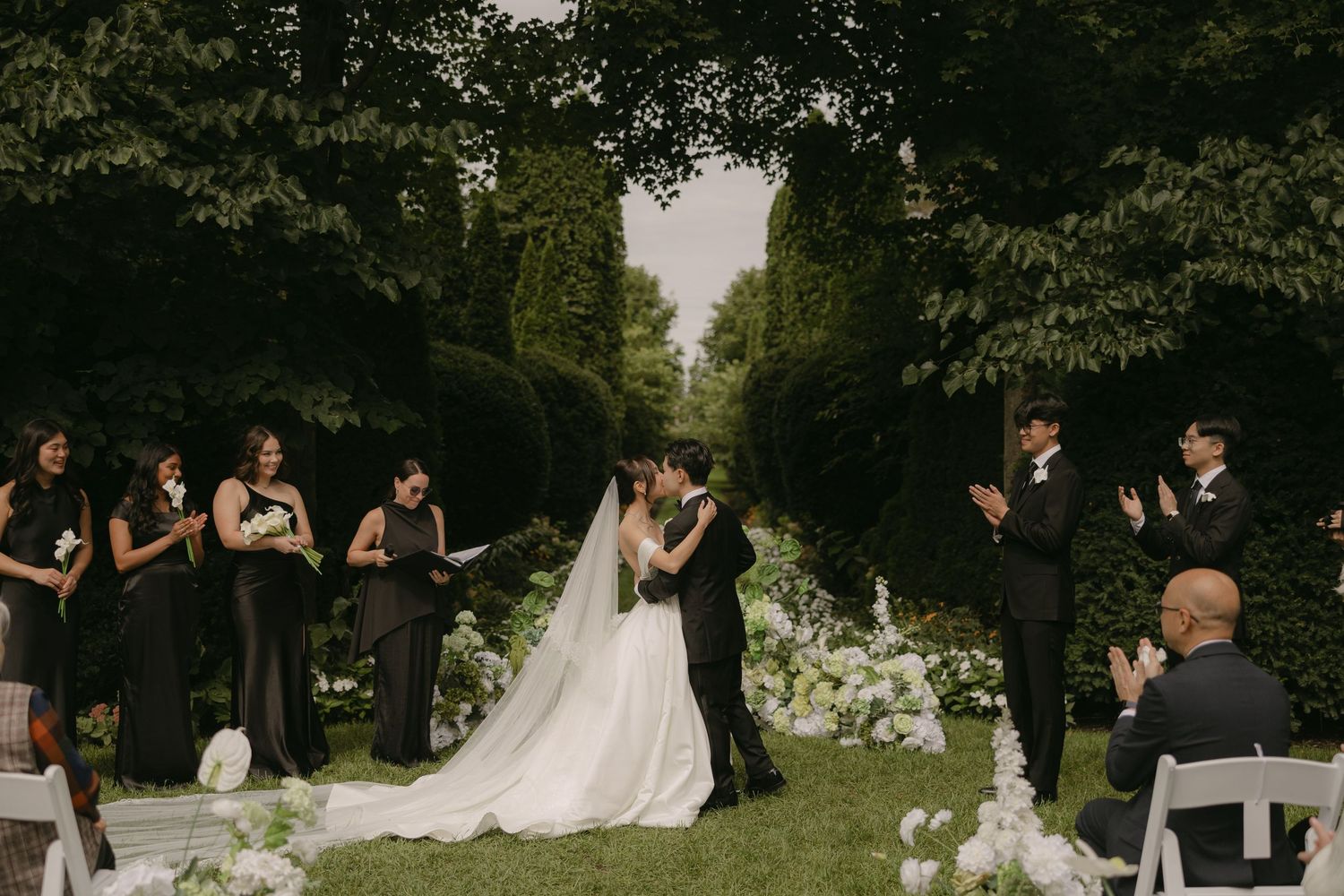 Bride and groom share first kiss surrounded by bridesmaids in lush garden ceremony.