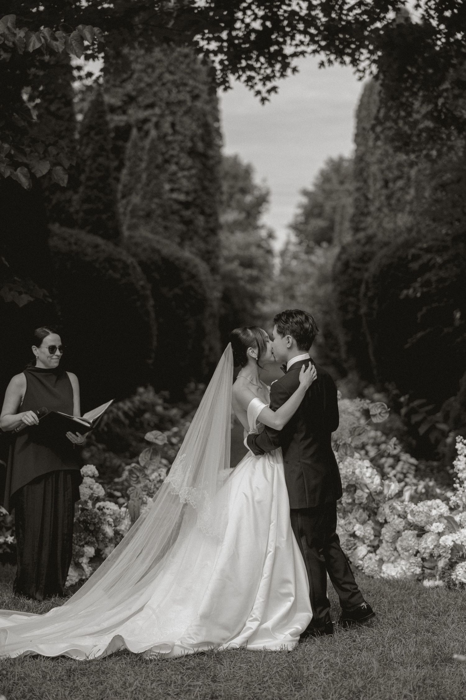 Black and white photo of bride and groom kissing during outdoor garden wedding ceremony.