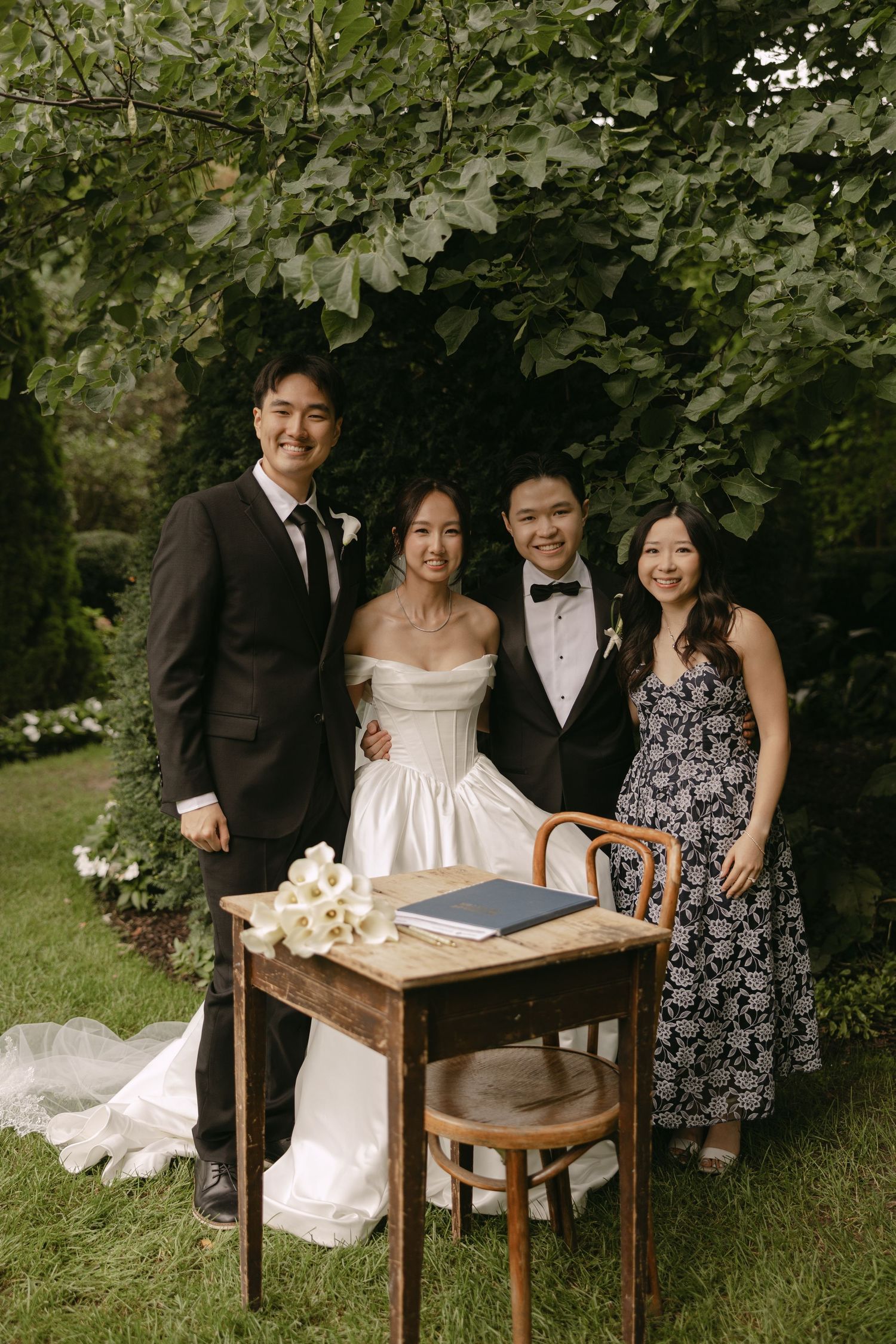 Newlyweds pose with two guests beside signing table under green leafy canopy.