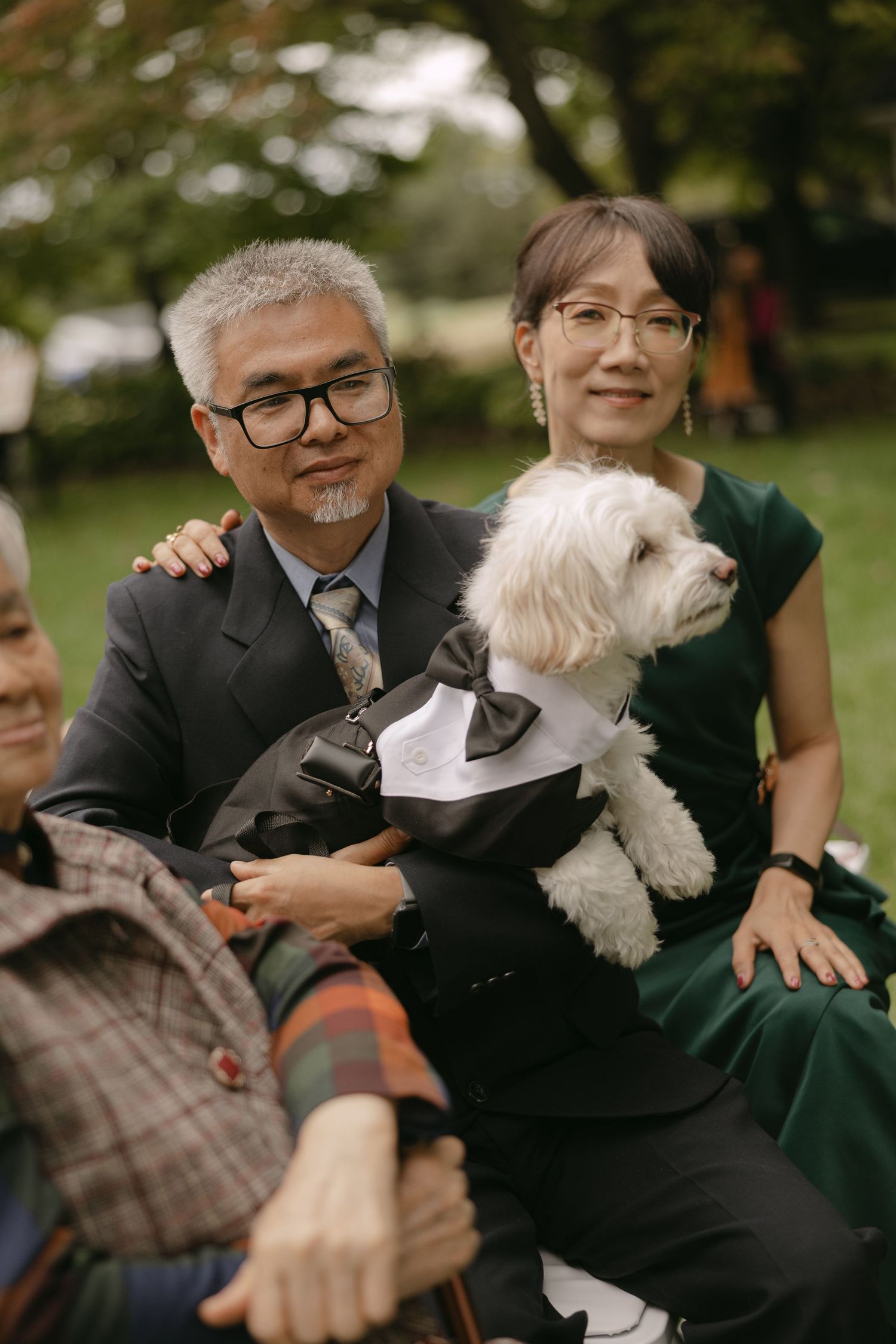 Older Asian couple in formal attire holding a white dog dressed in a tuxedo at an outdoor wedding.