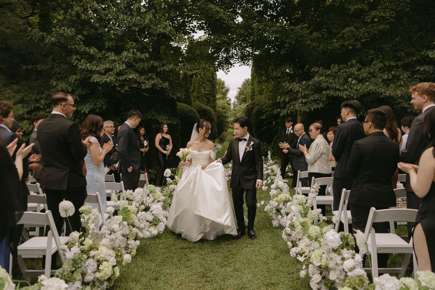 Bride and groom walk down a flower-lined aisle surrounded by guests at an outdoor garden wedding.