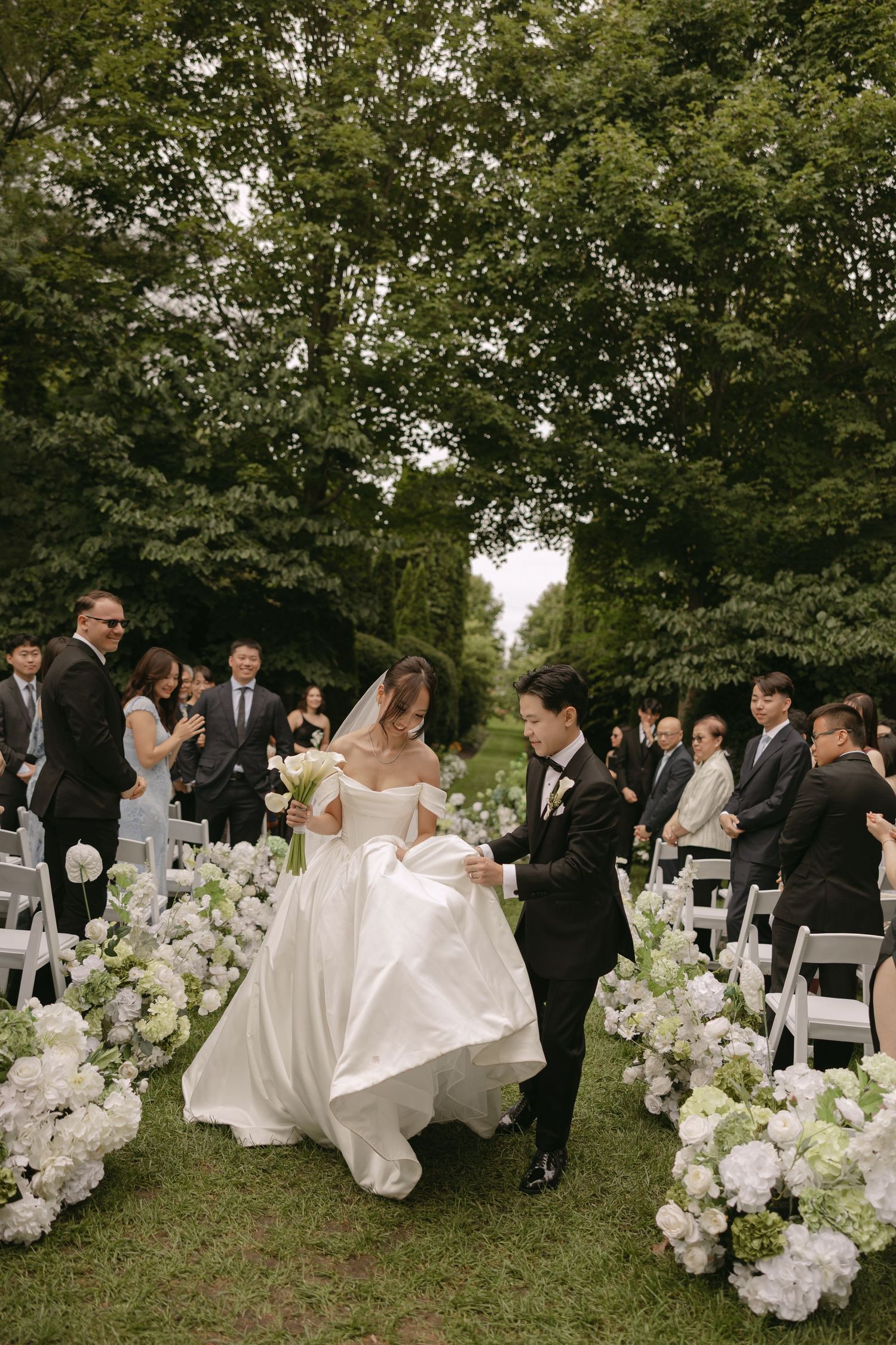 Groom helps bride lift her white gown while walking down a floral aisle at an outdoor wedding.