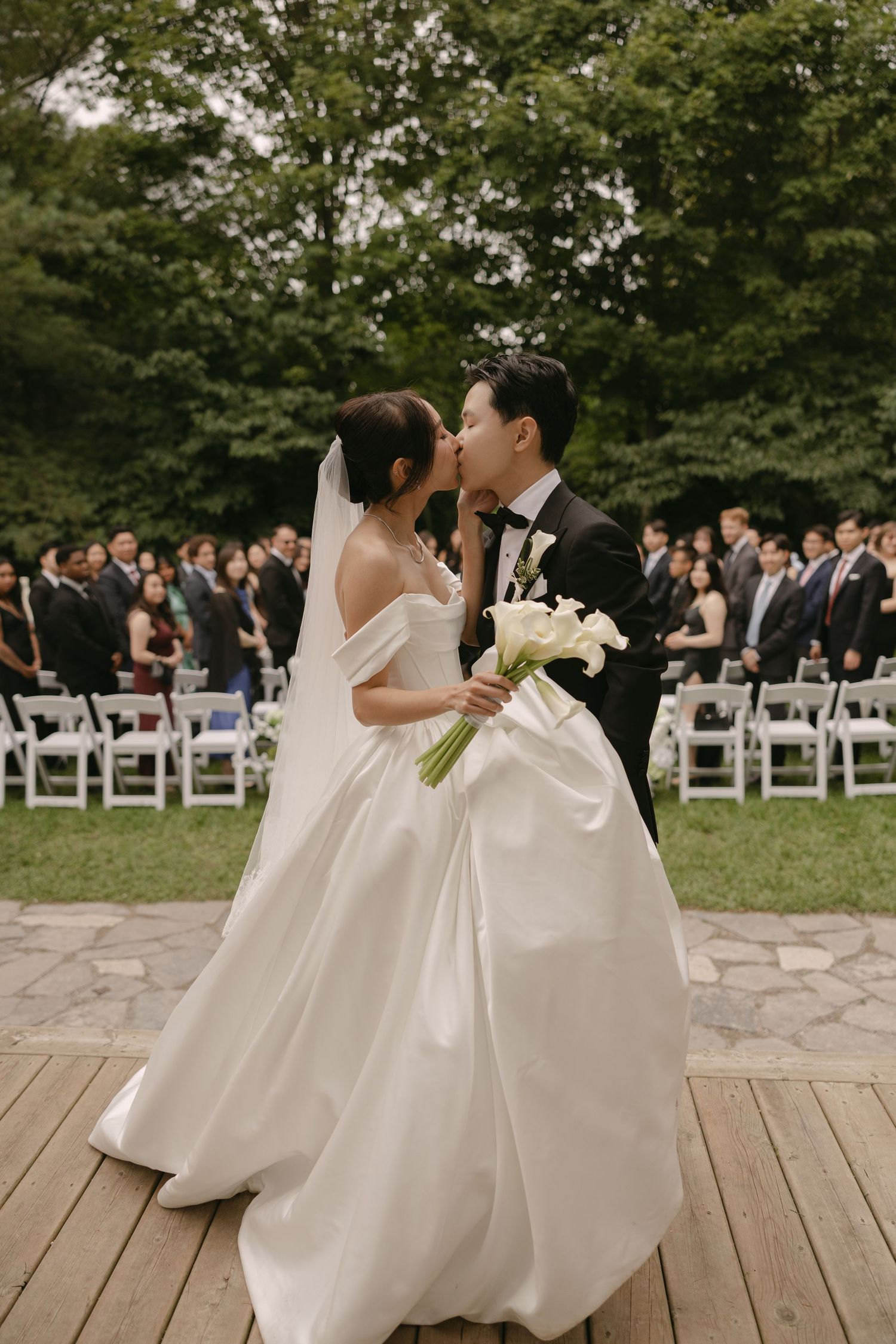 Bride and groom share a romantic kiss on a wooden deck with wedding guests seated in the background.