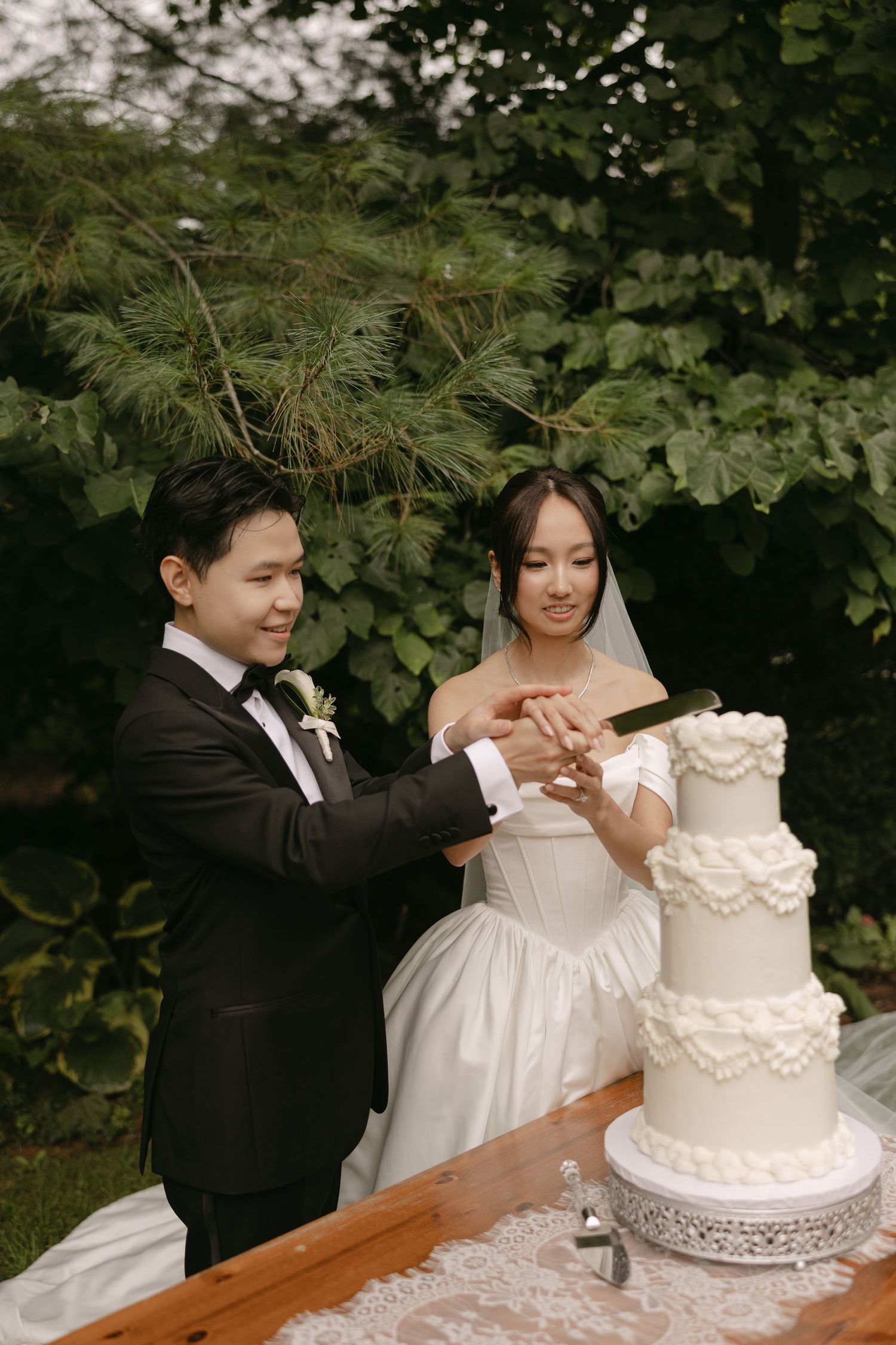 Asian bride and groom cutting a white multi-tiered wedding cake outdoors surrounded by greenery.