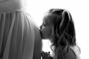 A young girl kissing a pregnant woman's belly in a tender black and white maternity photo.