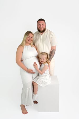 Pregnant woman in white dress poses with bearded husband and young blonde daughter in bright studio maternity photo.