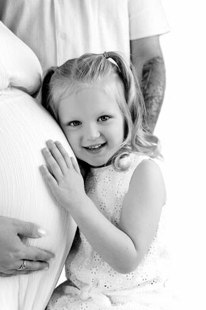 A smiling toddler girl hugs her mother's pregnant belly in a black and white maternity photo.