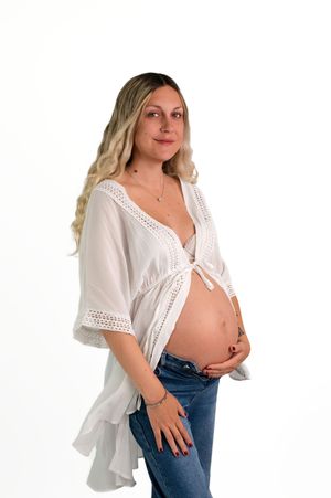 Pregnant woman in white lace top and jeans cradling her baby bump against a white background.