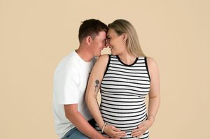 A couple sharing a tender moment, the woman visibly pregnant, posing together against a beige background.