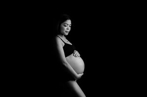 Black and white maternity photo of a pregnant woman cradling her baby bump against a dark background.