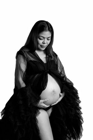 Black and white maternity photo of a pregnant woman in a sheer ruffled gown cradling her bare baby bump.