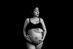 Black and white maternity photo of a tattooed pregnant woman in lingerie cradling her baby bump against dark background.