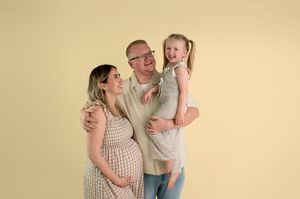 Pregnant woman in floral dress poses with smiling man holding young girl against yellow background.