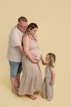 A pregnant woman in a gingham dress poses with her husband and young daughter against a soft yellow background.