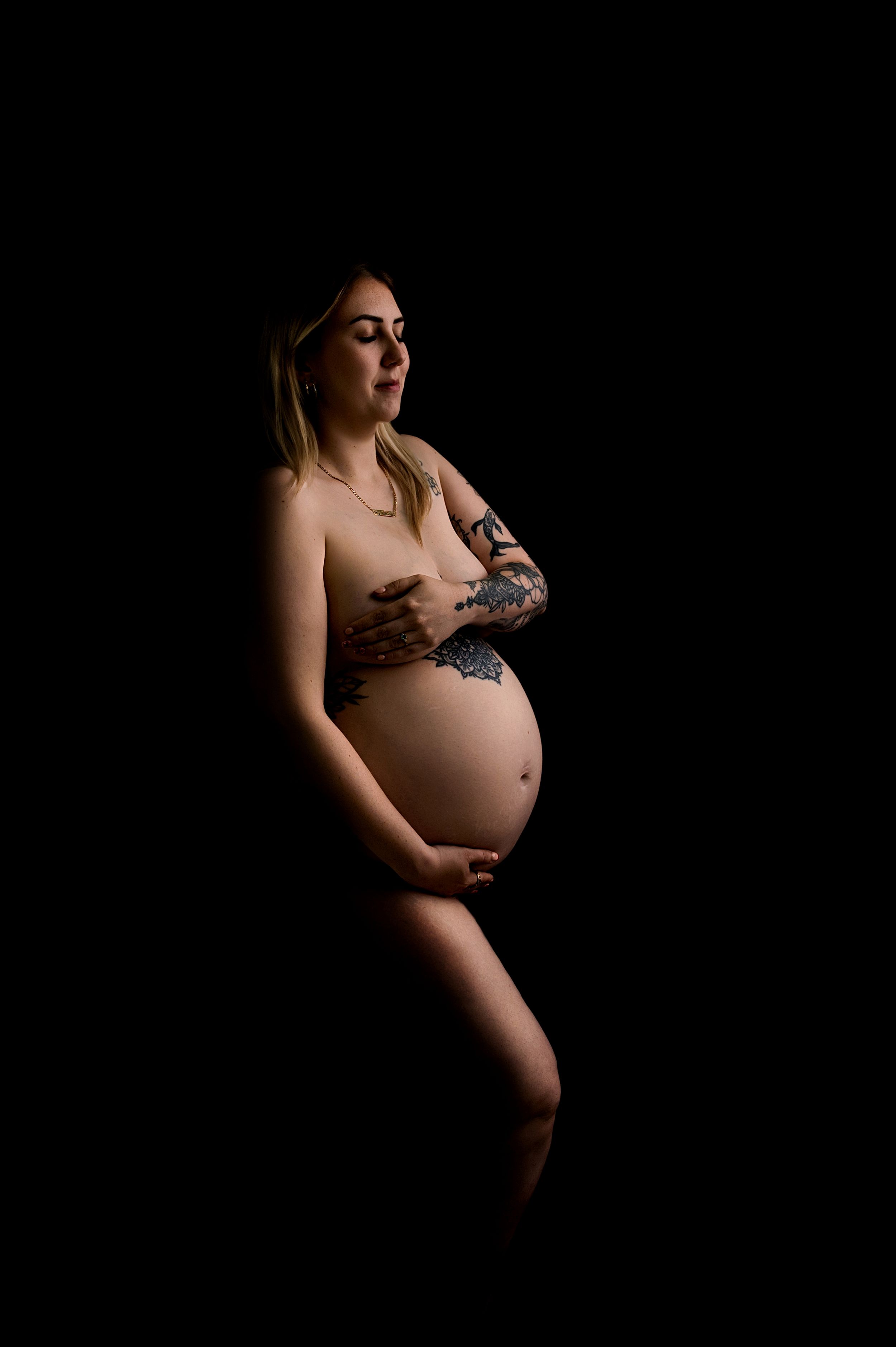 Pregnant woman with tattoos posing in a dark studio, cradling her baby bump in a maternity portrait.