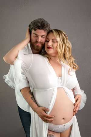 Couple sharing a tender maternity photo, man embracing pregnant woman in white robe, studio portrait.