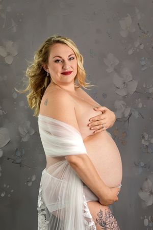 Pregnant woman in sheer white wrap poses gracefully against a floral gray backdrop in a maternity portrait.