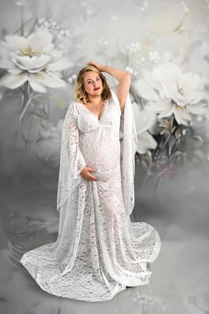 Pregnant woman in white lace gown with flowing sleeves poses gracefully against a floral backdrop.