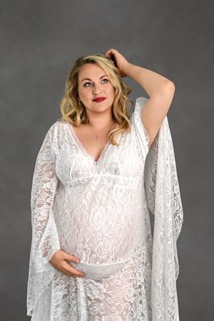 Pregnant woman in white lace maternity gown posing gracefully against gray studio background.