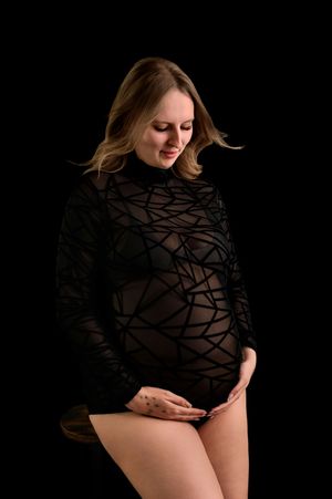 Pregnant woman in black sheer geometric bodysuit cradling her baby bump against a dark studio background.