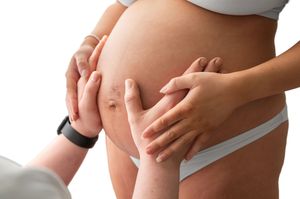 Pregnant woman's bare belly being gently touched by two pairs of hands, showing stretch marks on white background.