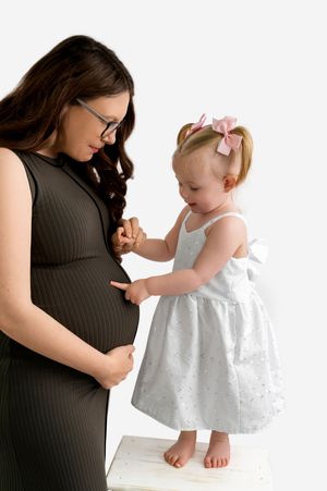 Pregnant woman in black dress with toddler girl in white dress touching her baby bump in studio.
