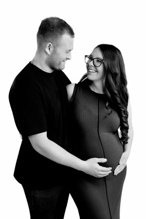 Black and white maternity photo of smiling couple, pregnant woman in ribbed dress with glasses, man in black t-shirt.