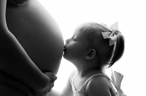Black and white photo of a toddler girl with a bow kissing a pregnant woman's belly in a tender maternity moment.