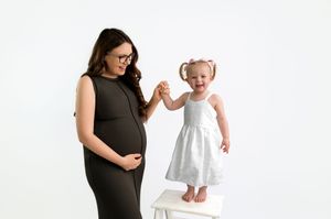 Pregnant woman in black dress holds hands with toddler girl in white dress standing on stool, white background.