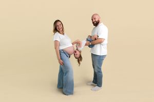 Pregnant woman and bearded man playfully hold a child upside down, posing together for a fun family maternity photo.