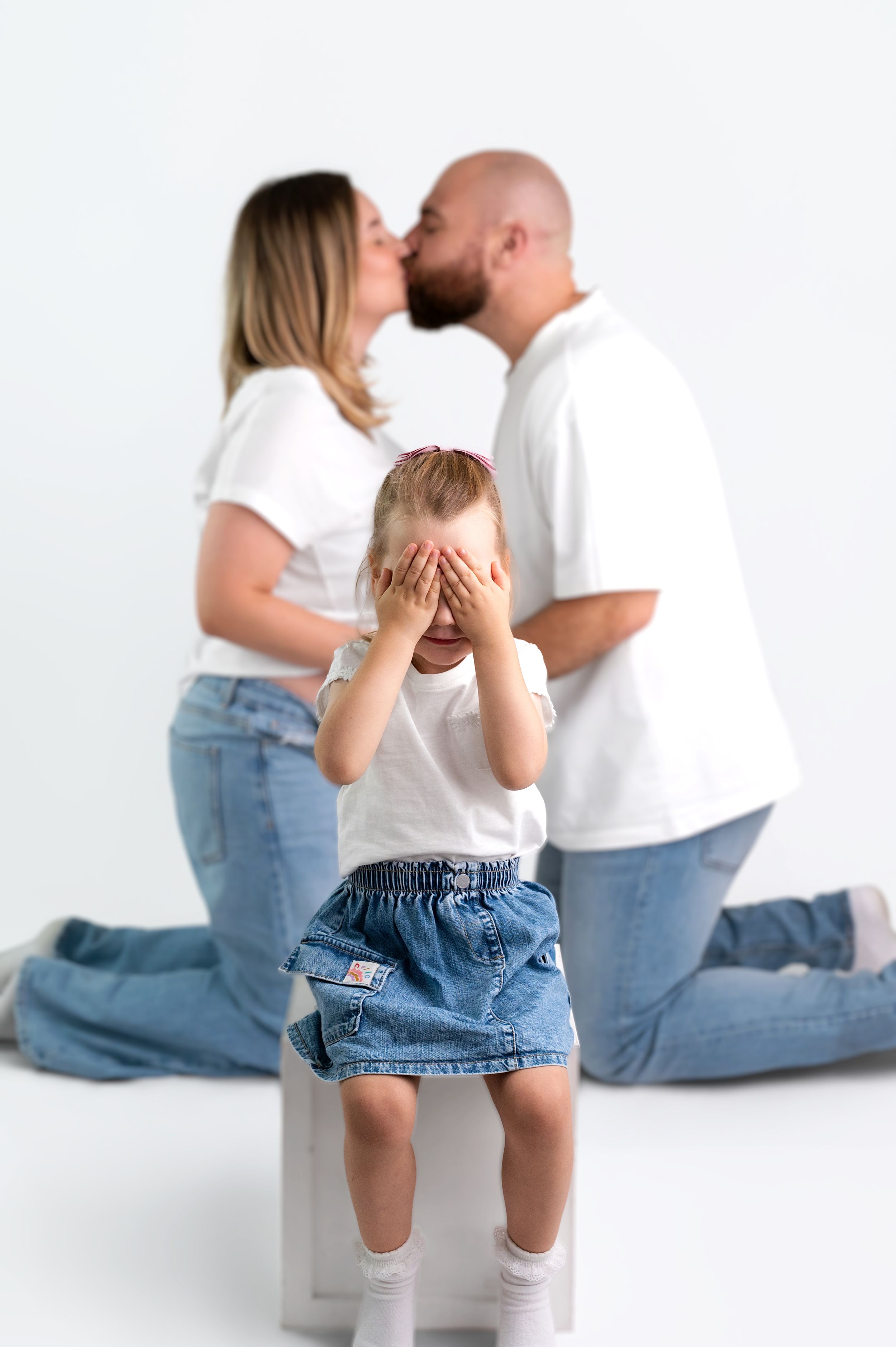 A young child covers her eyes as her parents share a kiss behind her in a bright white studio setting.