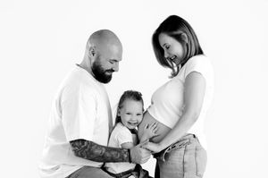 Black and white maternity photo of a bald tattooed man, pregnant woman, and toddler smiling together.