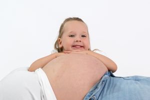 Young girl resting her chin on her pregnant mother's baby bump, smiling happily against white background.