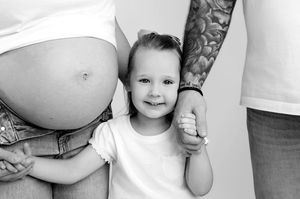 Black and white photo of a pregnant woman and tattooed man holding hands with a smiling toddler girl between them.