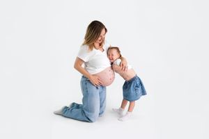 Pregnant woman kneeling on white background with toddler girl kissing her baby bump.