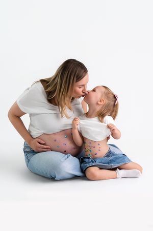 Pregnant mother kissing toddler daughter, both wearing jeans with colorful stickers on their bellies, on white background.