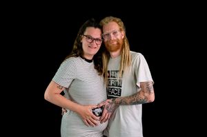Pregnant couple posing together, woman in striped dress and man with red dreadlocks and tattoos, against black background.