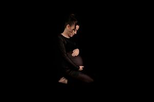 Pregnant woman in black outfit sits on dark background, cradling her baby bump in a serene maternity photo.