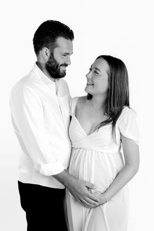 Black and white maternity photo of smiling couple in white outfits, man embracing pregnant woman's belly.