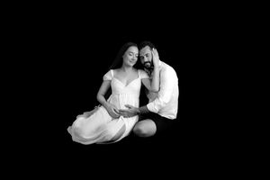 Black and white maternity photo of a couple sitting on floor, woman in white dress, man tenderly resting head on hers.