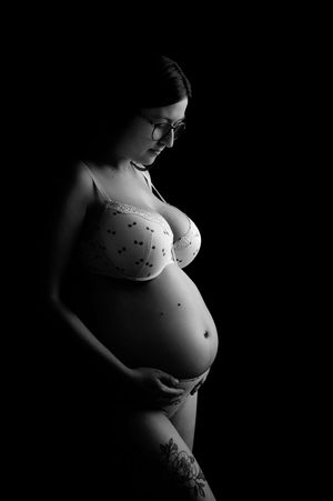 Black and white maternity photo of pregnant woman in lingerie with tattoo, cradling baby bump against dark background.