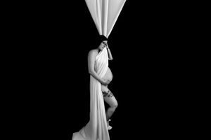 Black and white maternity photo of pregnant woman draped in white fabric against dark background.