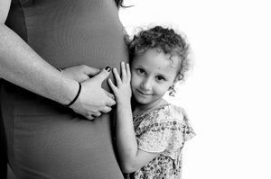 Black and white photo of a young girl hugging a pregnant woman's belly, smiling warmly at the camera.