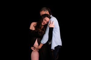 A couple embraces tenderly against a dark background, the woman in a black dress leaning into the man in a blue shirt.