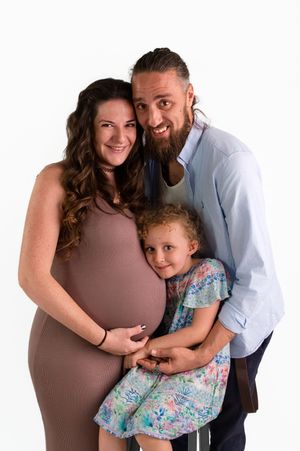 A pregnant woman in a brown dress poses with her bearded partner and young daughter in a studio maternity photo.