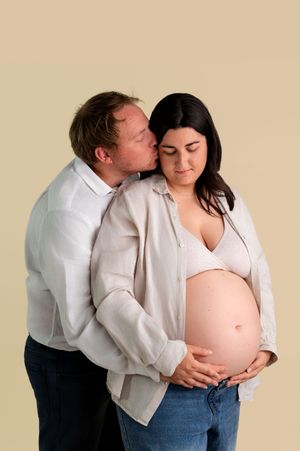 A man kisses a pregnant woman's cheek while cradling her baby bump in a tender maternity photo.