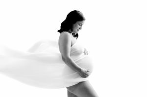 Black and white maternity photo of a pregnant woman in flowing white fabric against a white background.