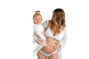 Pregnant woman in white lingerie holding smiling toddler girl against white background.