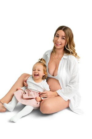 Pregnant woman in white shirt sitting with laughing toddler girl in pink dress on white background.