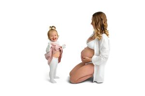 Pregnant woman kneeling beside laughing toddler girl in pink outfit on white background.